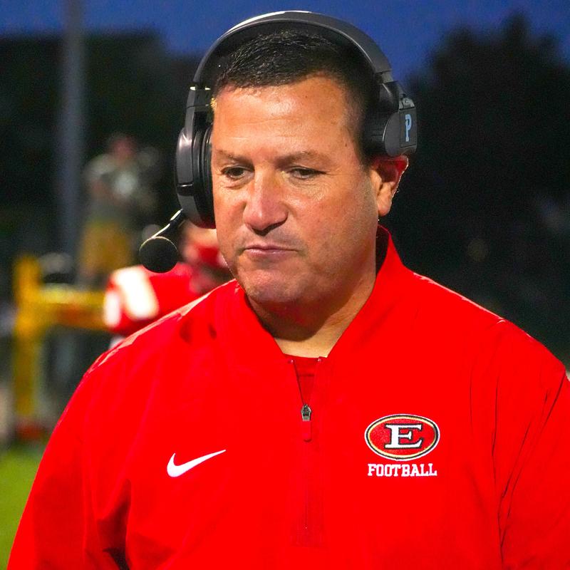 A man in a red jacket with a headset looks pensive on a sports field.