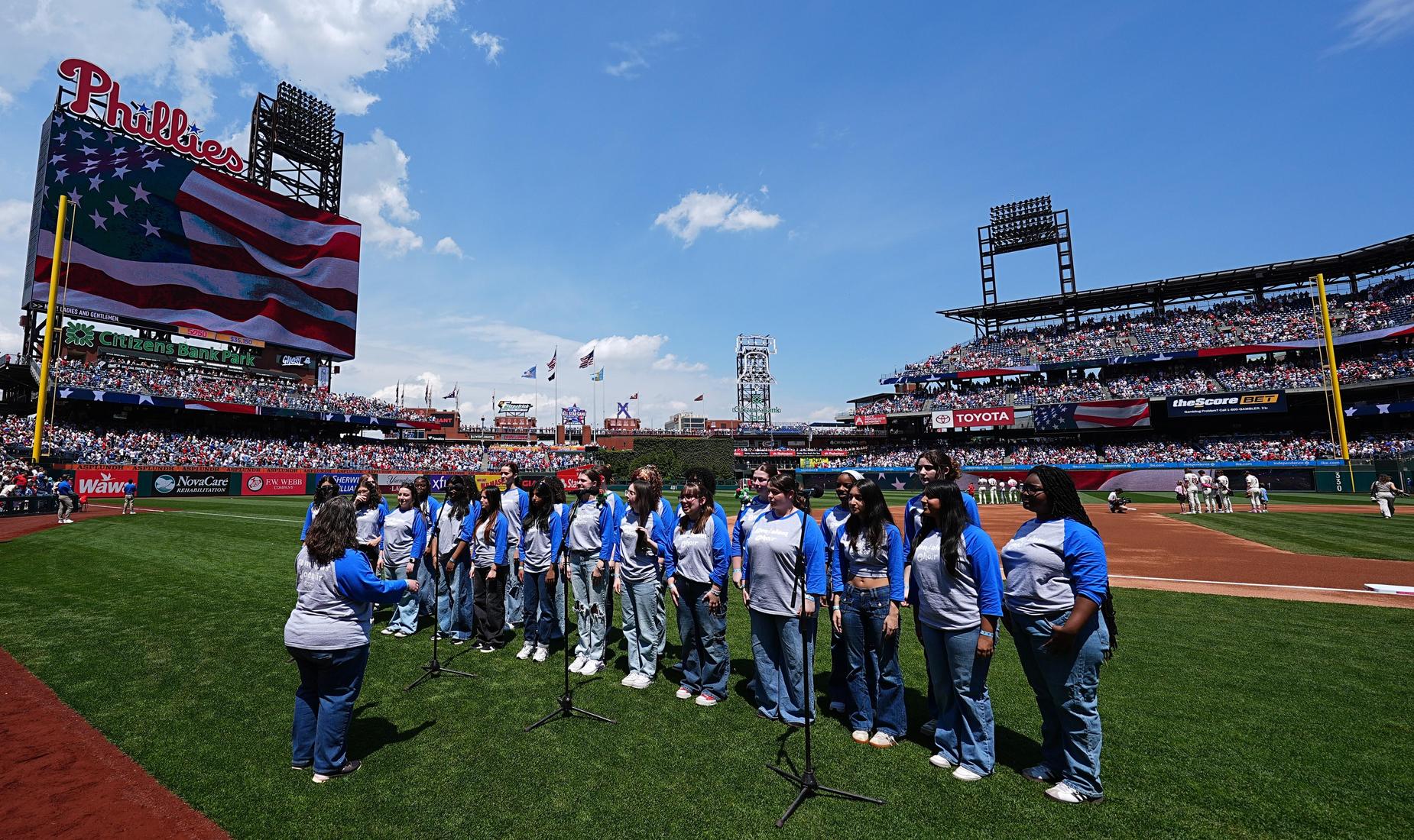 A choir performs in front of a large American flag at a baseball stadium.