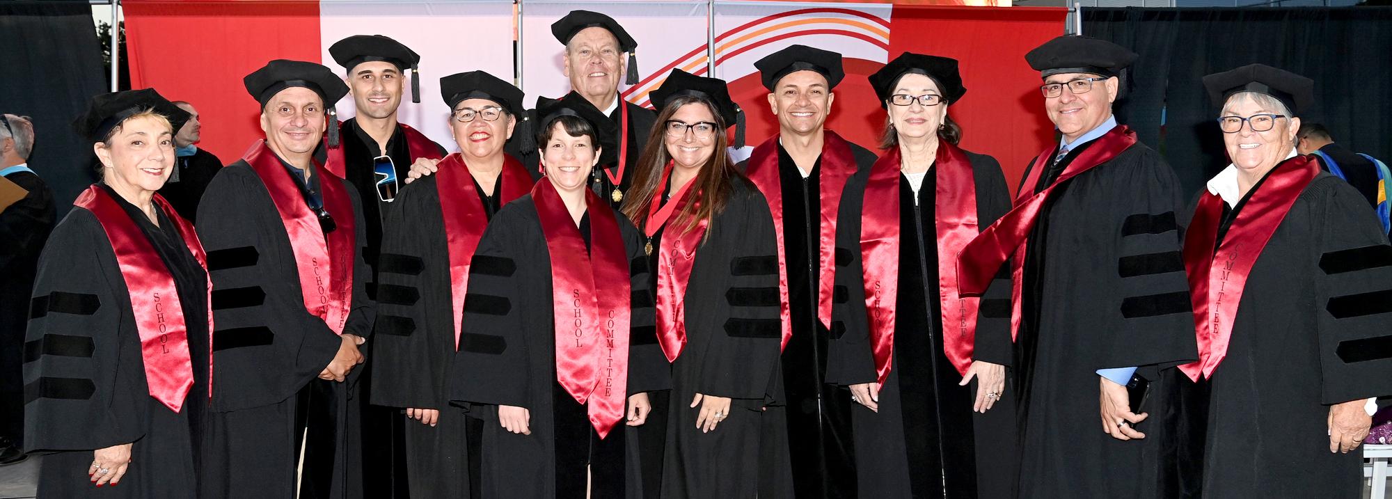 Group of graduates in caps and gowns with red sashes at a ceremony.