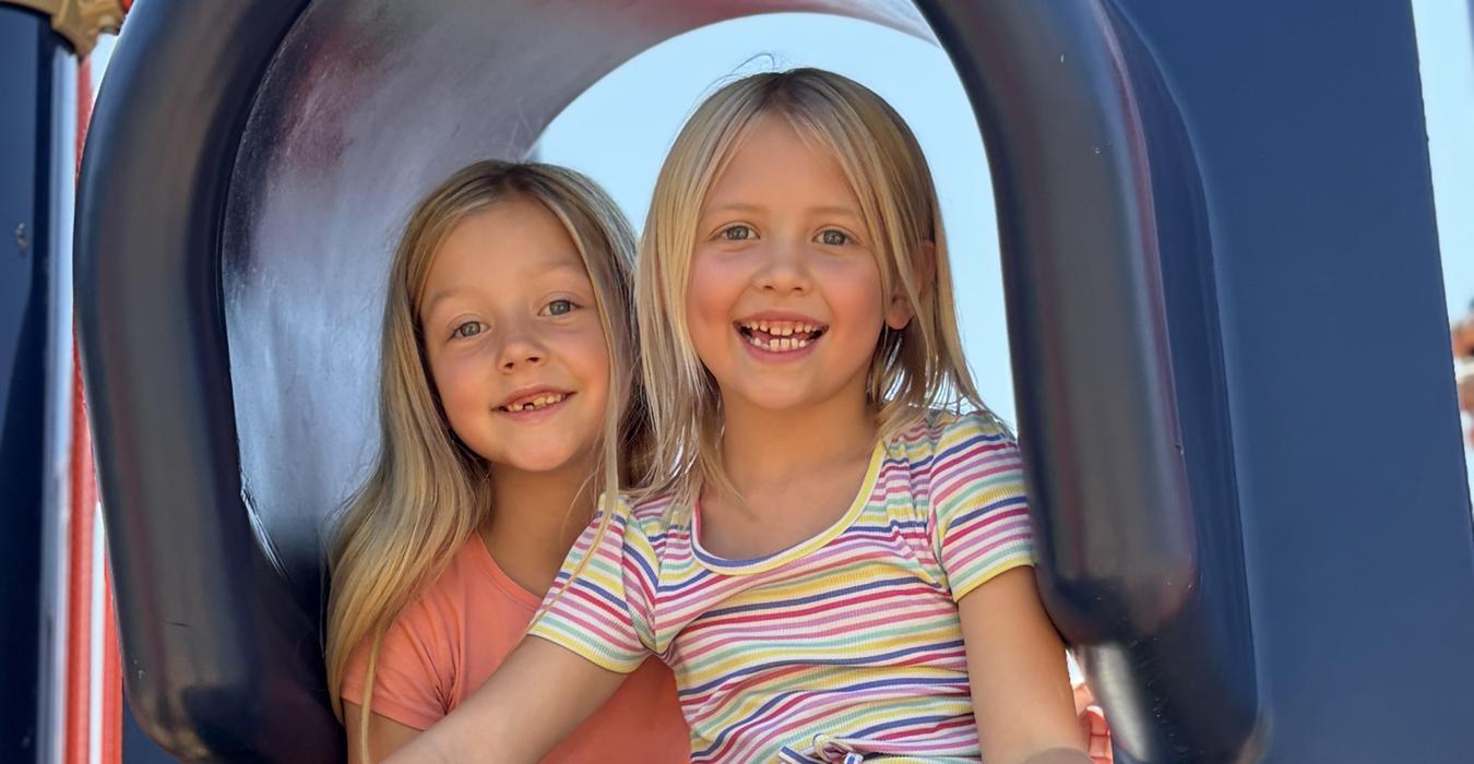 Two children sitting on a slide, smiling in bright clothes.