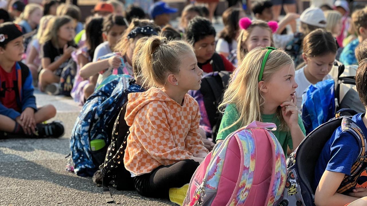 A group of children sitting on the ground, gathered for an event, with backpacks around them.