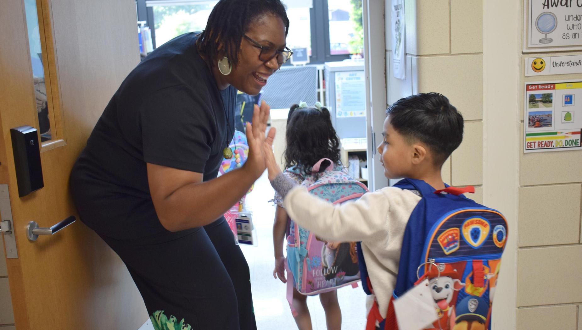 Teacher giving a high five to a child with a backpack at a classroom door.