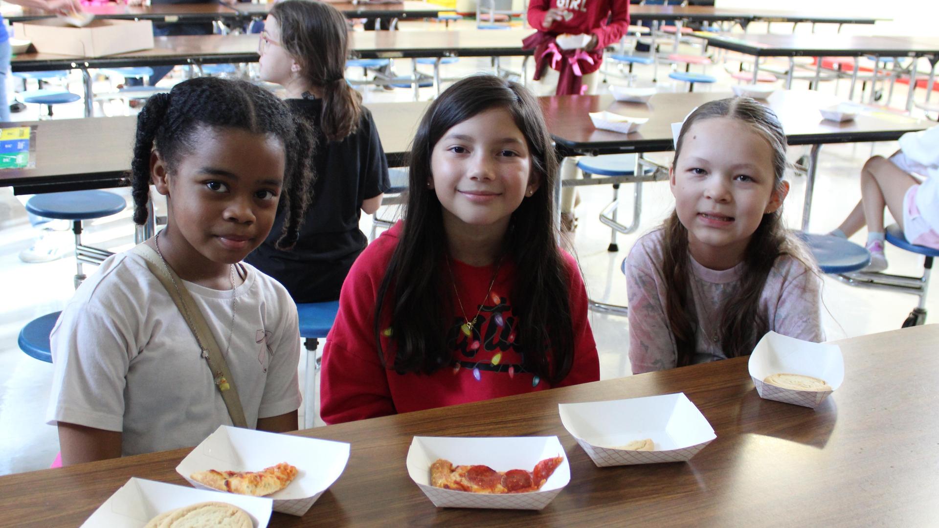 Three girls smiling at a table with snacks in a school cafeteria.