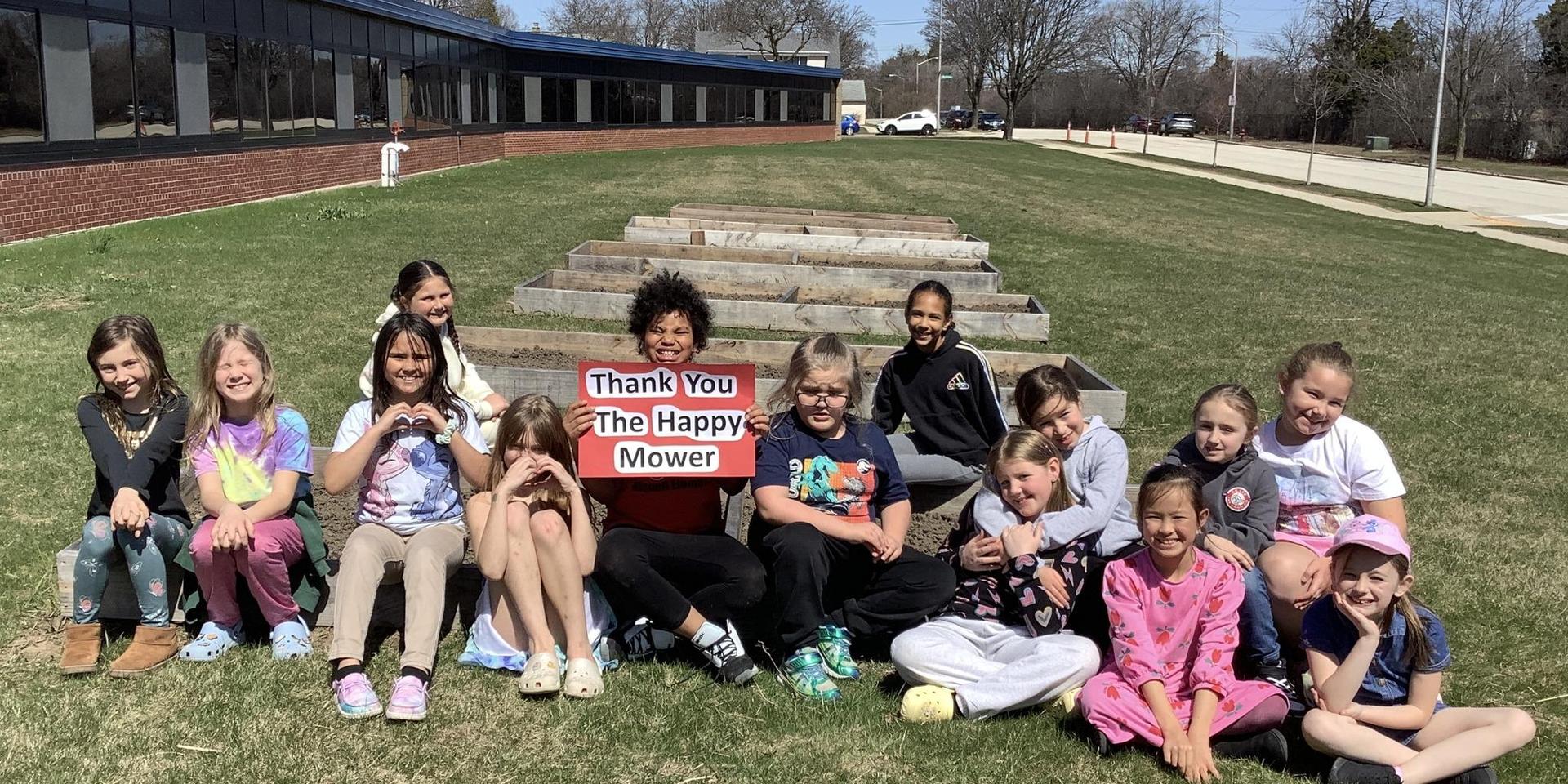 Group of children sitting outdoors with a thank you sign in a garden setting.