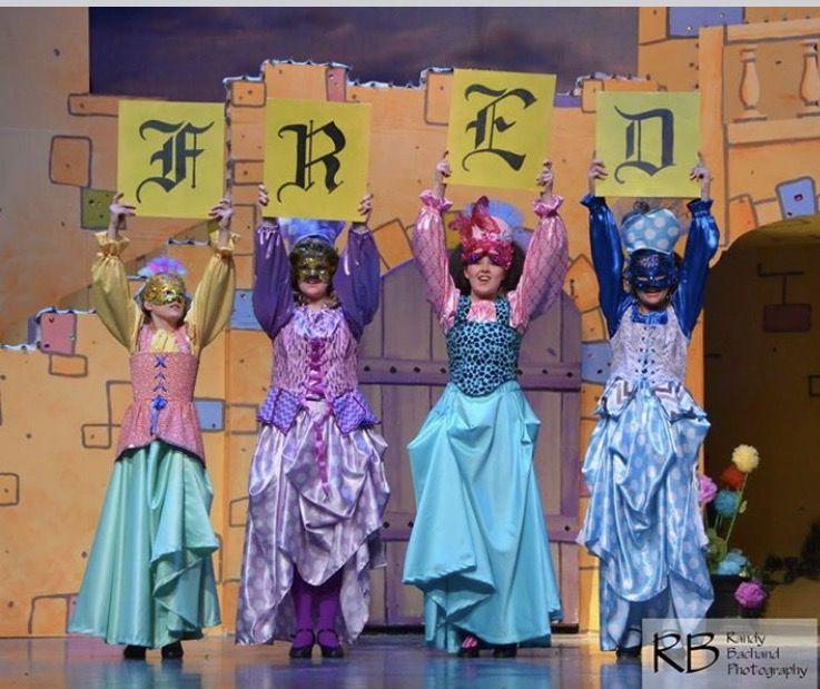 Four high school theater students in a play wear princess dresses and hold letters spelling 