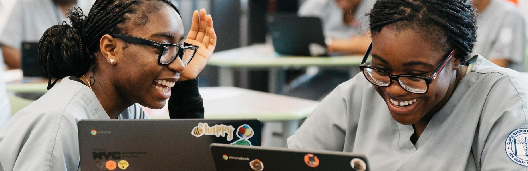 Students wearing gray scrubs are seated at desks in a classroom, working on laptops. The laptops have various stickers, including one with the NYC logo and another with colorful circular designs. A notebook with red binding is placed on the desk in front of one student. The classroom has green chairs and tables, and other students with laptops are visible in the background. Bright natural light enters through windows, creating a well-lit environment.