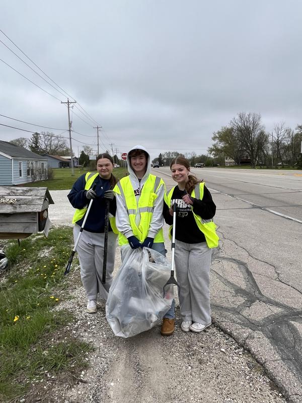 Three young volunteers posing with a trash bag on the roadside.