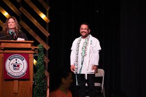 Male graduate wearing a white uniform top and a decorative green “Phlebotomy 2025” sash smiles brightly while standing near the podium on stage.