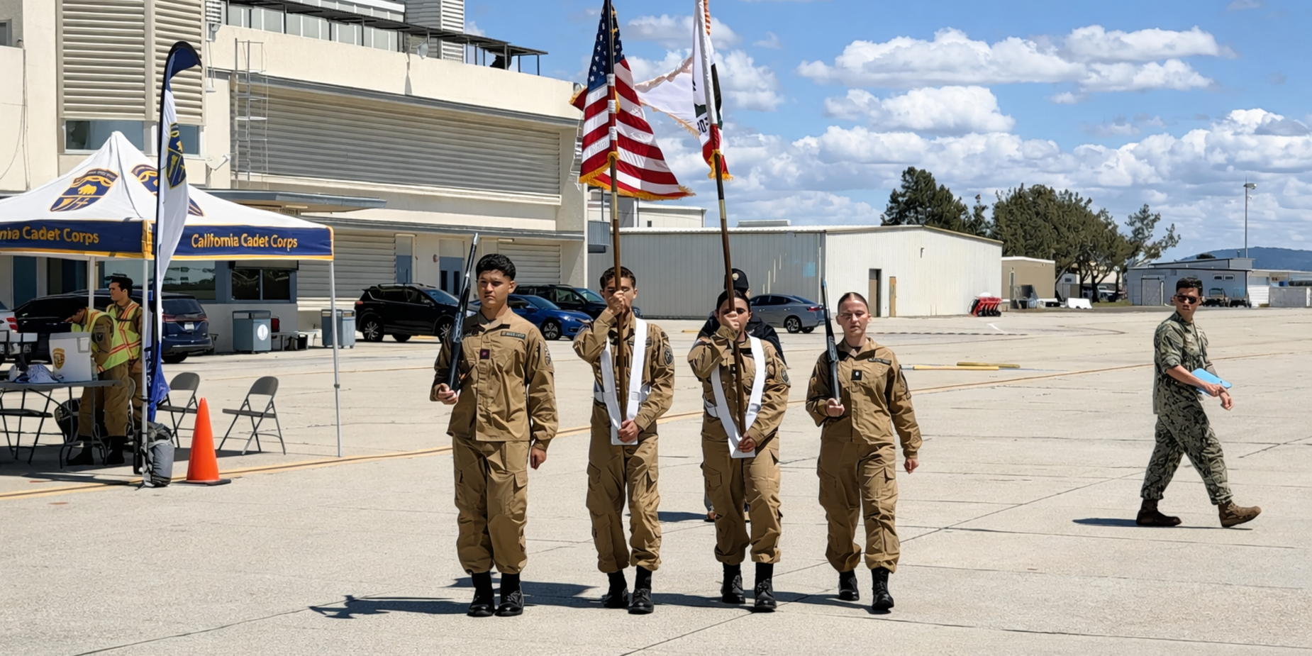 Four cadets in uniform carrying flags during a ceremony outdoors.