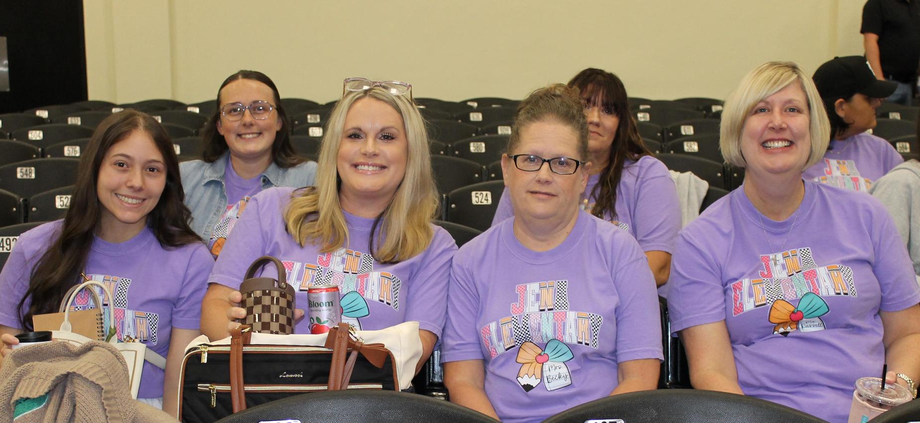 A group of five women wearing matching purple shirts, sitting in an auditorium.