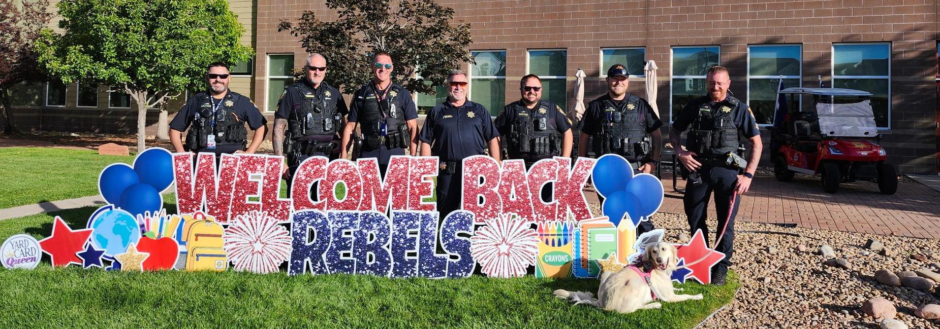 Law enforcement officers standing in front of a building with a welcome back sign.