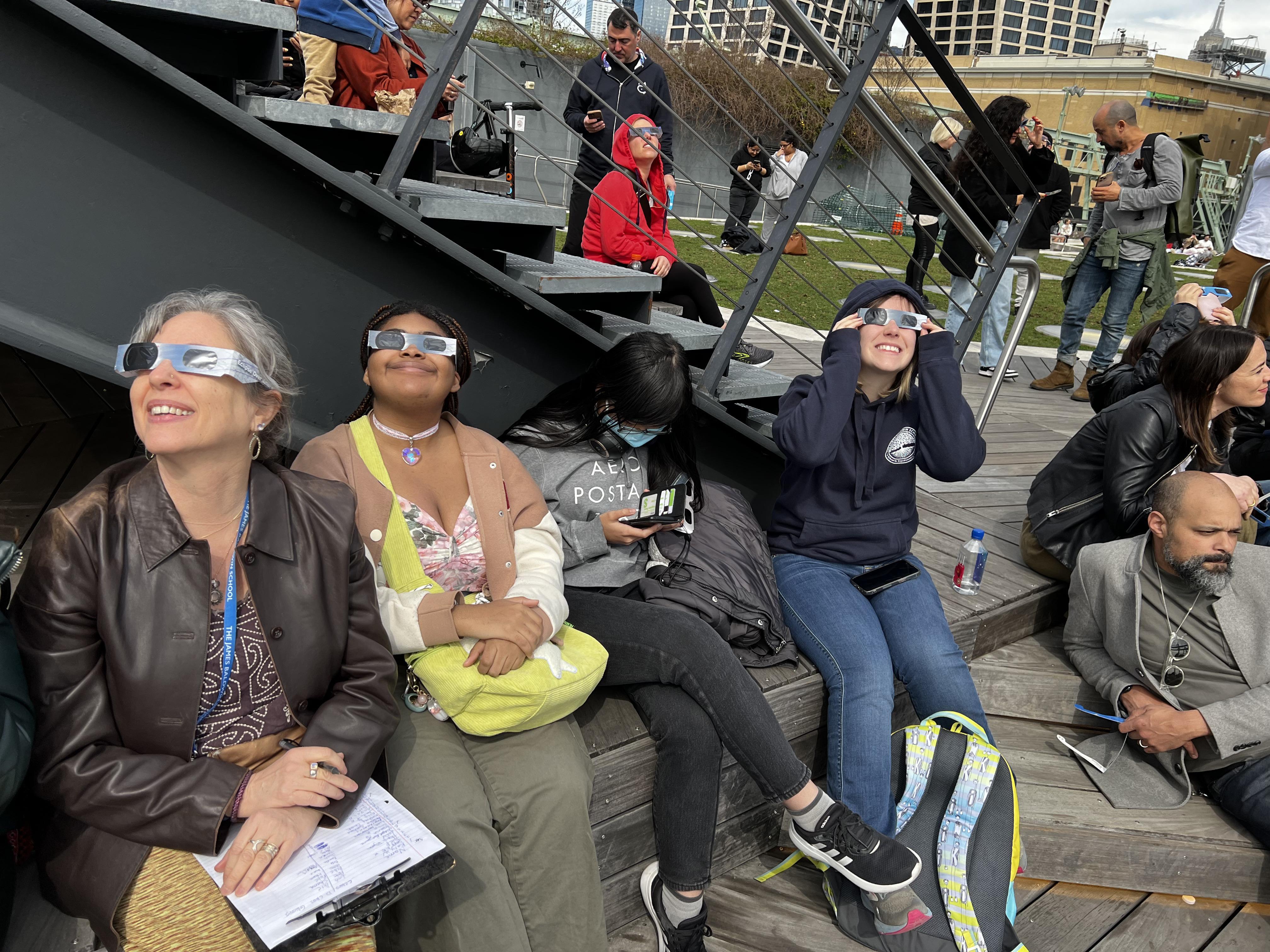 Four people wearing solar eclipse glasses sit on a bench and look up at the Solar Eclipse