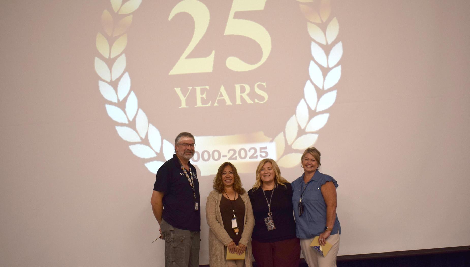 Four people pose in front of a backdrop celebrating 25 years, with a golden laurel design.