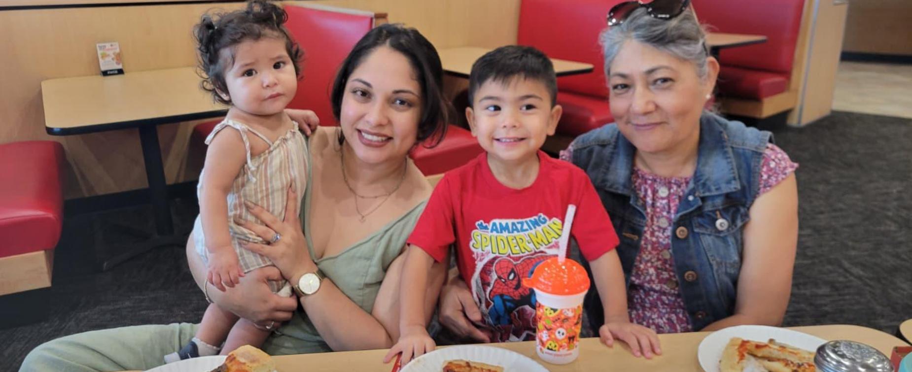 A woman holds a toddler while sitting with a young boy and an older woman at a restaurant booth.