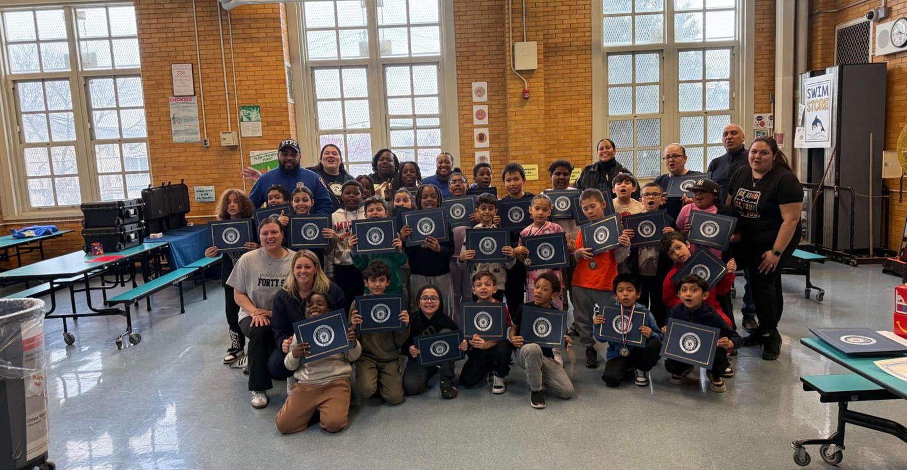 A group of children and adults holding certificates in a school cafeteria.