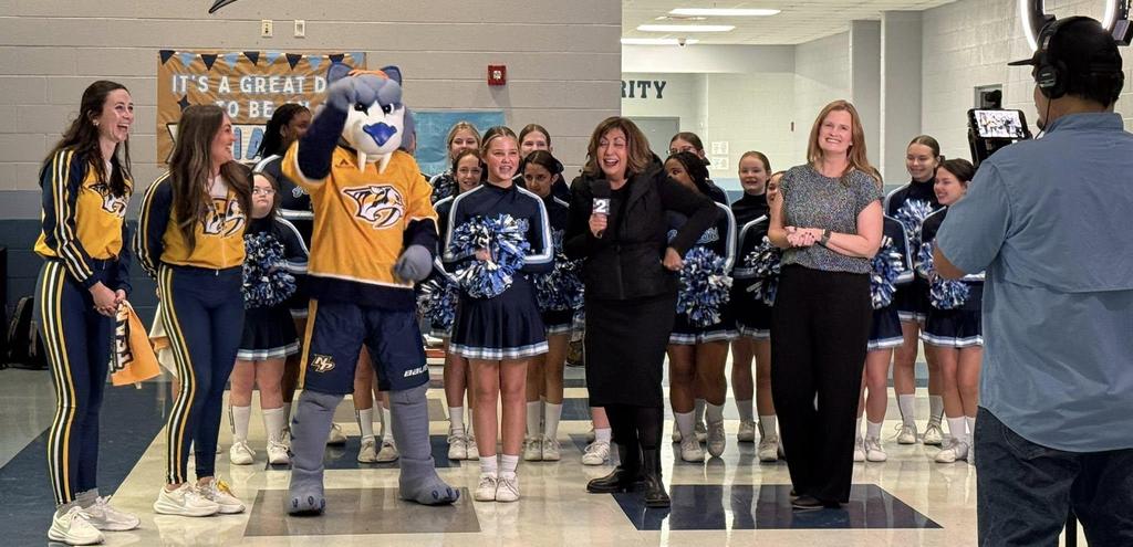 cheerleaders from middle school and from nashville predators hockey team along with the mascot stand cheering with 2 adults posing for a camera man and holding a microphone