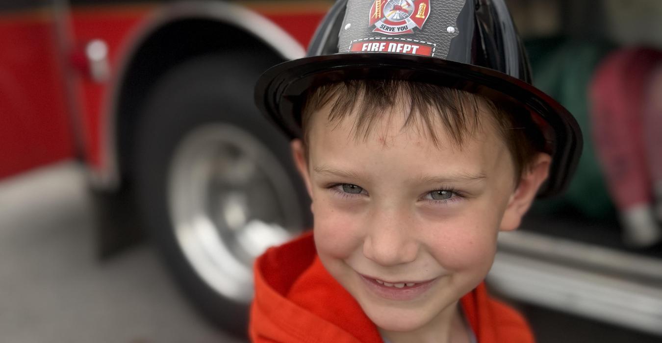 Young boy wearing a black fire department hat and smiling in front of a fire truck.
