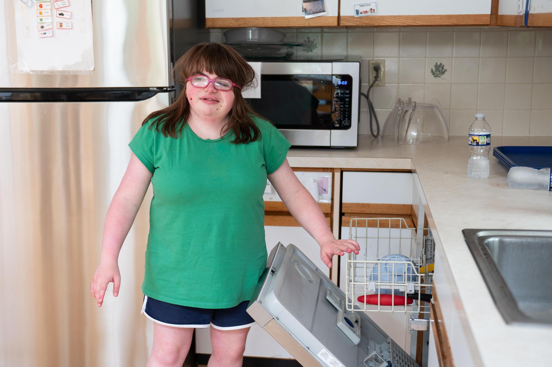 Girl in green shirt and glasses closing dishwsher.