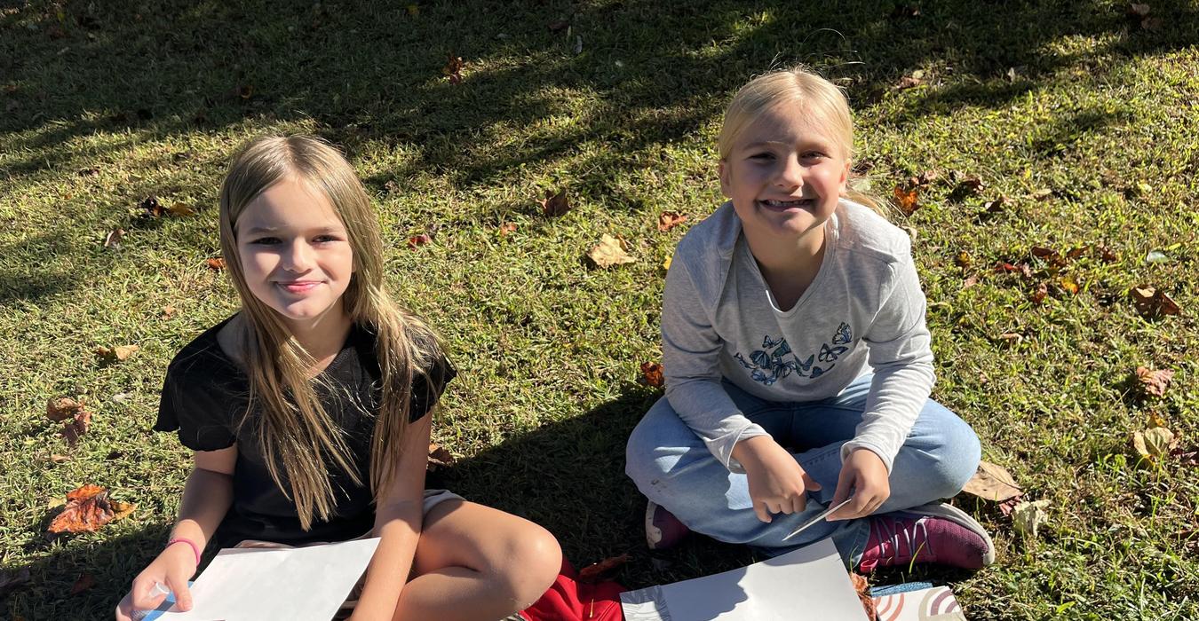 Two girls sitting outdoors on grass, smiling and holding blank paper.