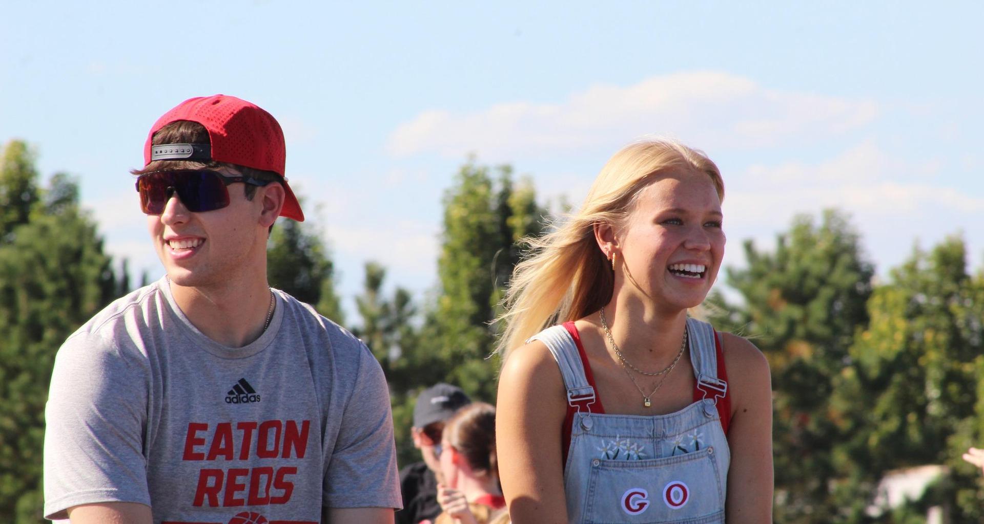 Two smiling students riding in a parade, surrounded by greenery.
