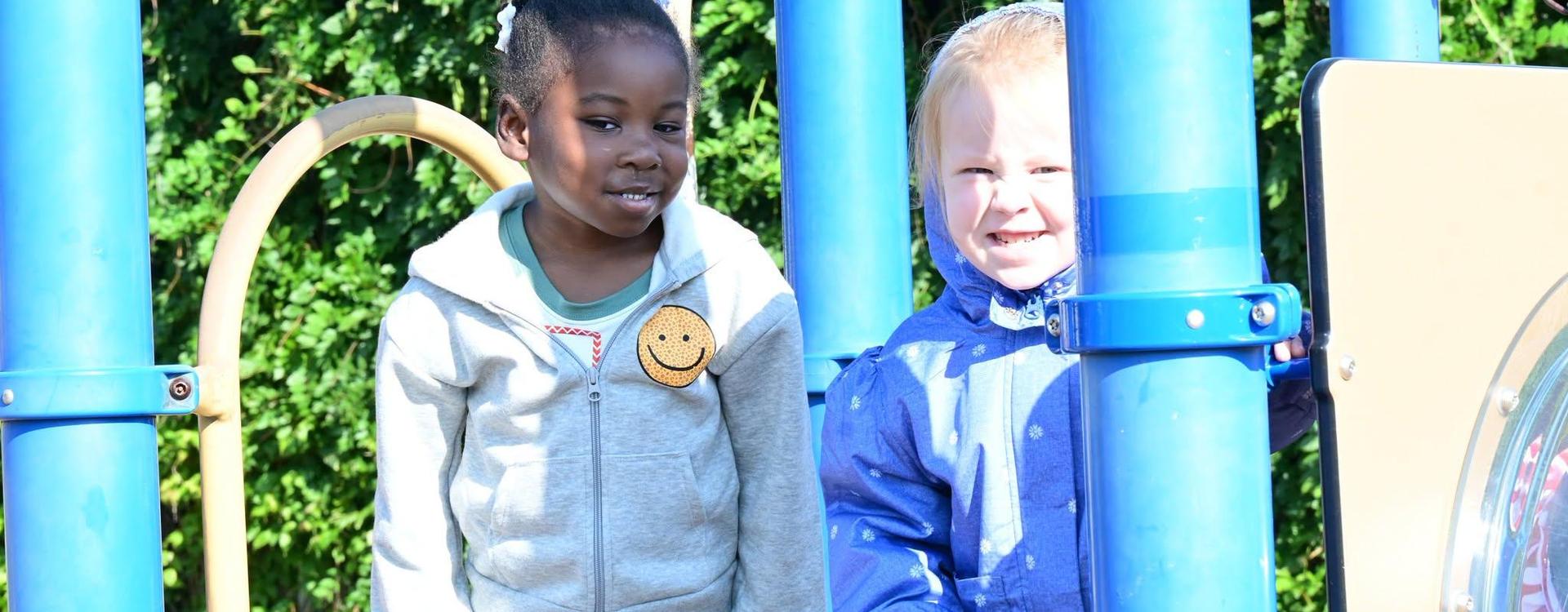 Two children play on a playground, one smiling and the other looking serious.