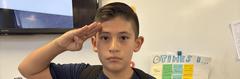 A boy saluting in a classroom setting with a chart in the background.