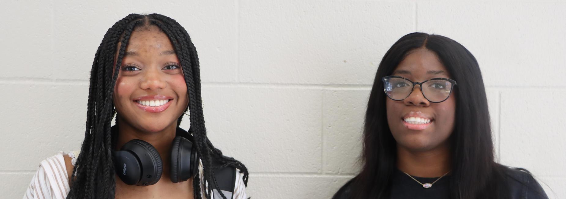 Two young women smiling at the camera, standing against a light gray wall.