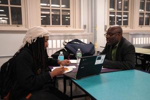 Two people discussing at a table with laptops and drinks in a study area.