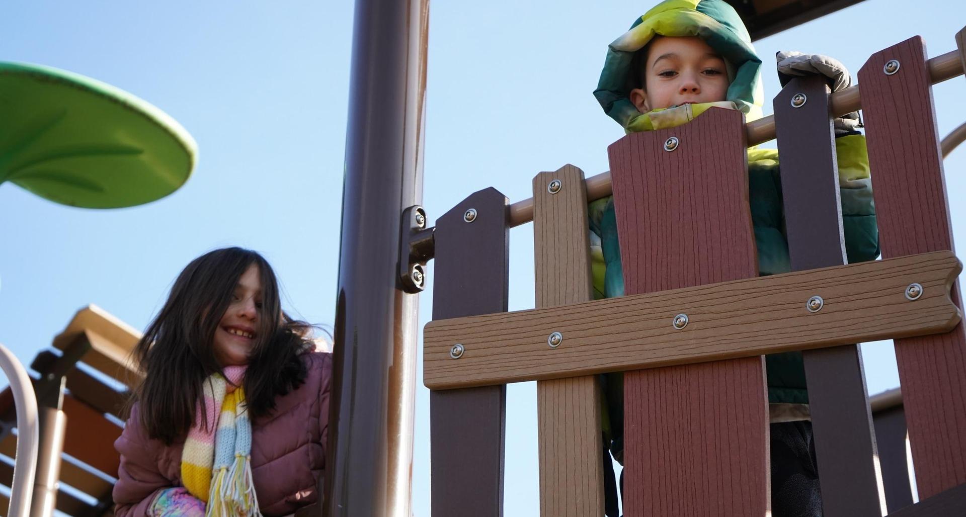 A boy and girl play on a playground, one peeking from behind a fence.