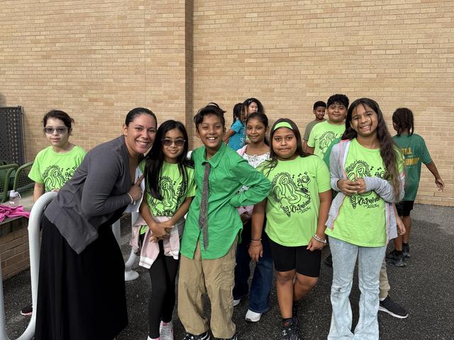 A group of children in matching green shirts pose with an adult outside a building.