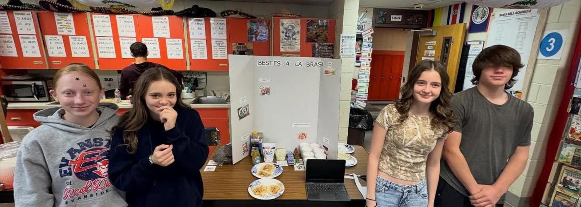 Four students pose with a project board and food in a classroom.