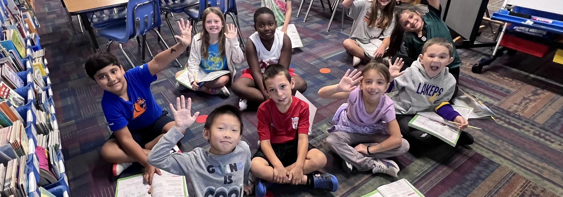 Students sitting on the floor in a classroom, smiling and waving at the camera.