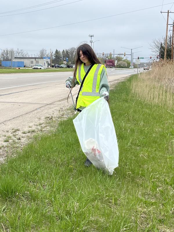 A volunteer in a neon vest collecting litter near a roadside grass area.