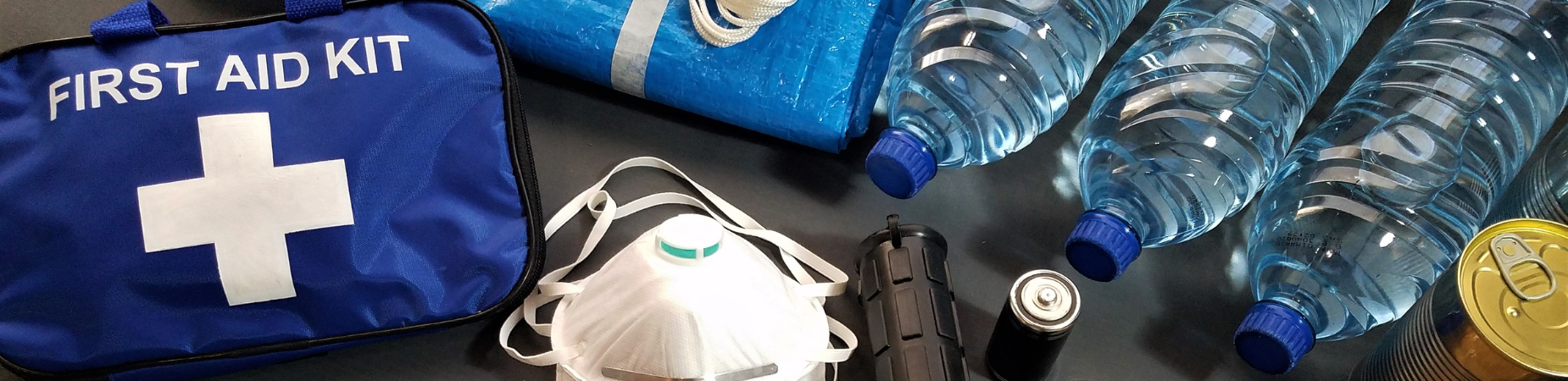 A first aid kit, bottled water, a mask, flashlight, and a can on a dark surface.
