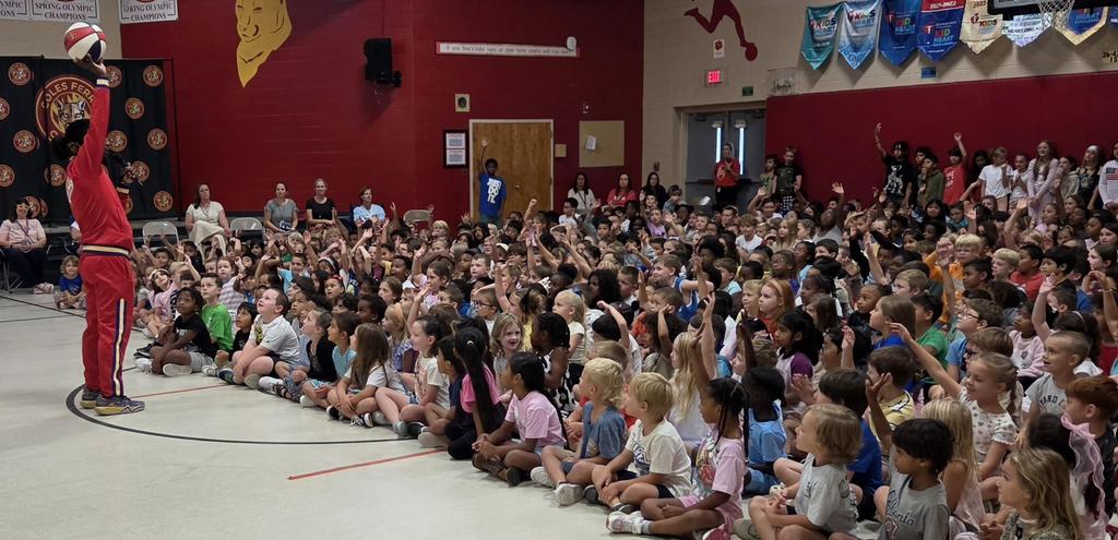 A performer entertains a large group of children seated on the gym floor.