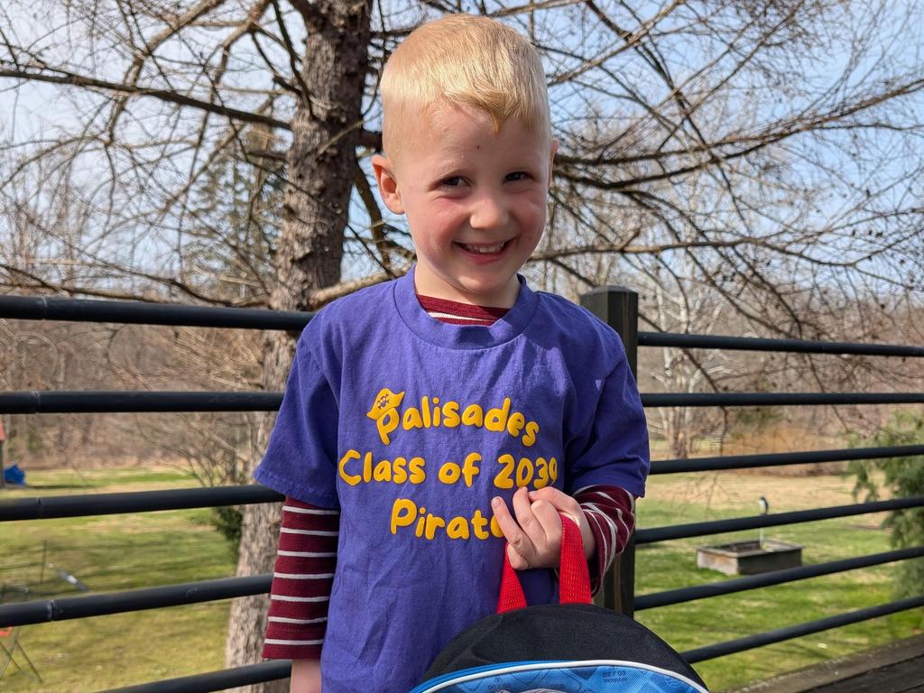 An incoming kindergarten boy holding a backpack and wearing a "Palisades Class of 2039 Pirates" purple t-shirt.