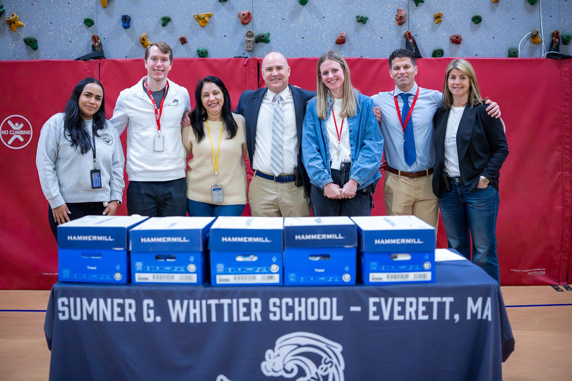 A group of six individuals poses behind a table with boxes in a school gym.