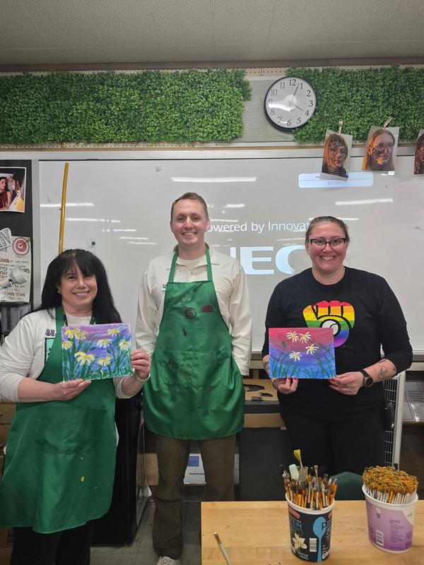 Three individuals showcasing their paintings in a classroom setting.