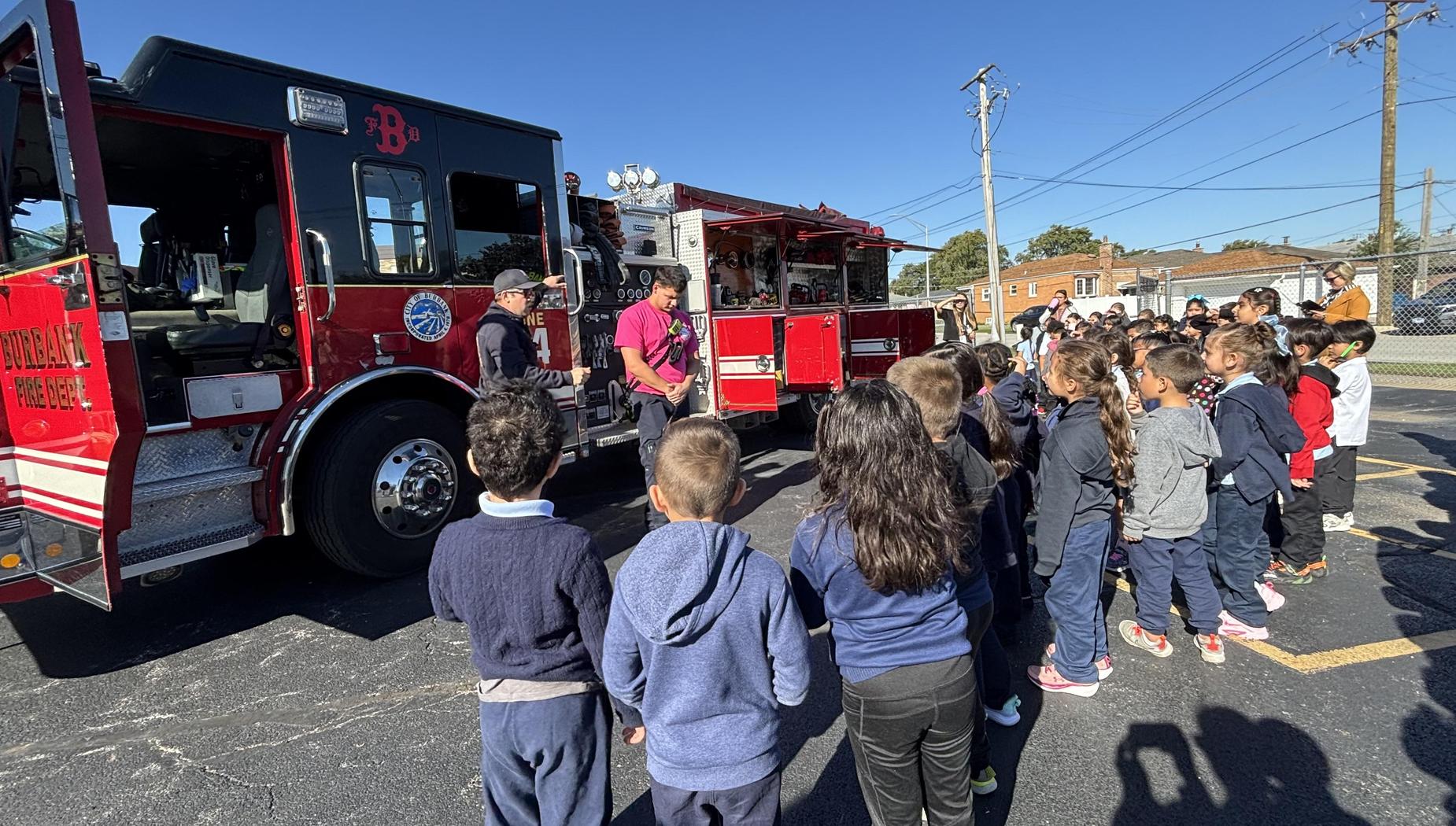 Children gather in front of a fire truck while firefighters explain safety.
