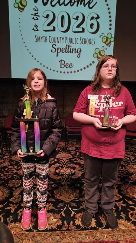 two students standing on a stage holding Spelling Bee trophies after the county spelling bee