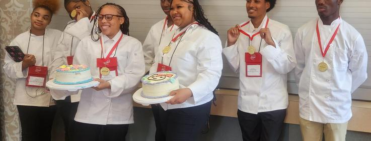 Six individuals in chef uniforms, smiling and holding decorated cakes at an event.