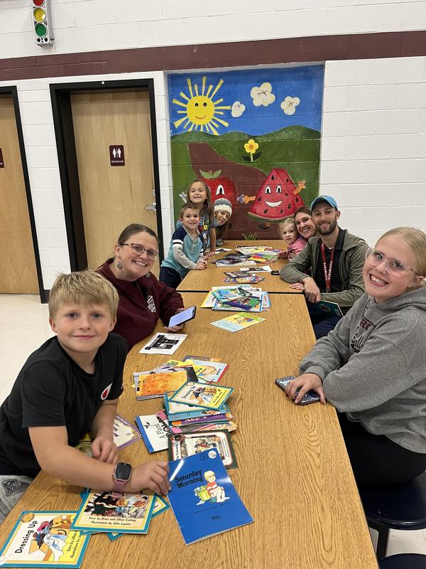 A group of children and adults gather at a table with books and artwork in the background.