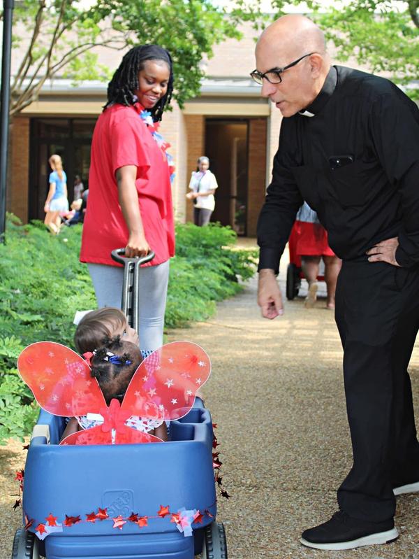 A priest interacts with a child in a wagon, guided by a woman.