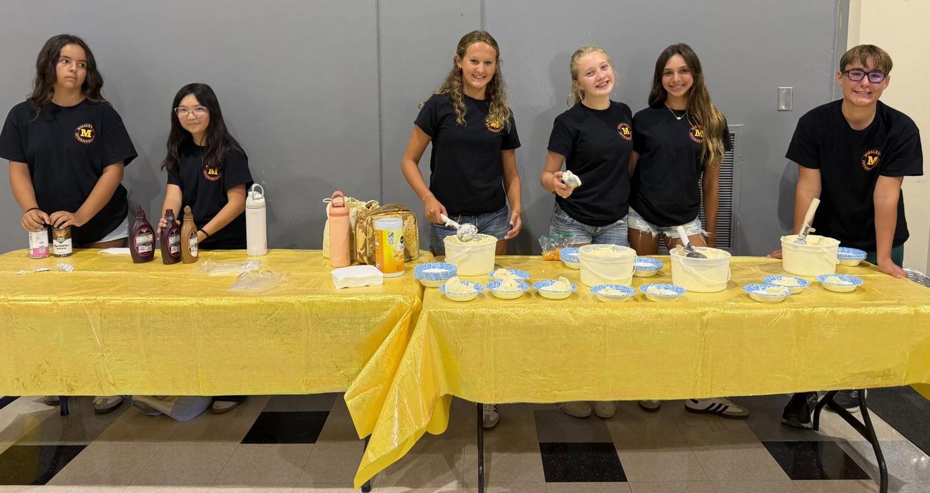 Six young people preparing food at a table with condiments and toppings.