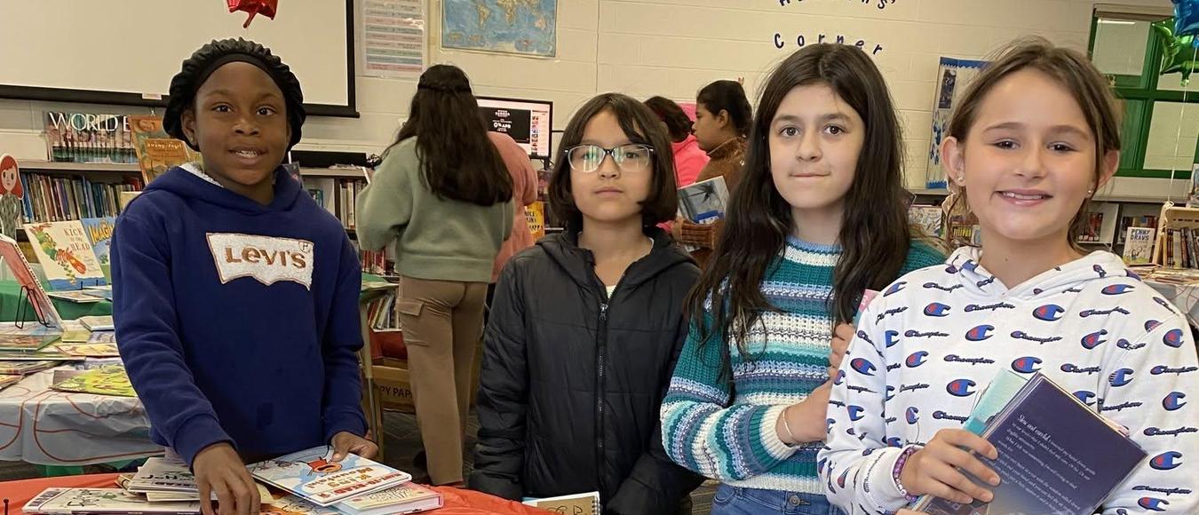 Four young students smile by a round table covered in children's books inside a school library.