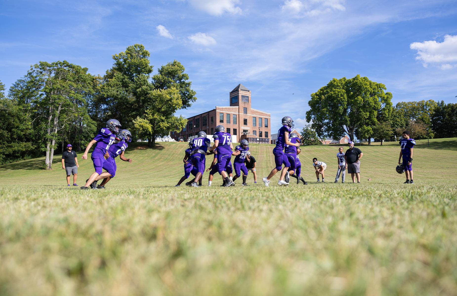 Youth football players practicing on a sunny field with a school building in the background.