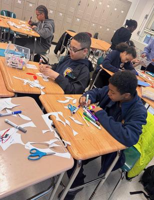 Students engaged in a craft activity at desks with scissors and paper cutouts.