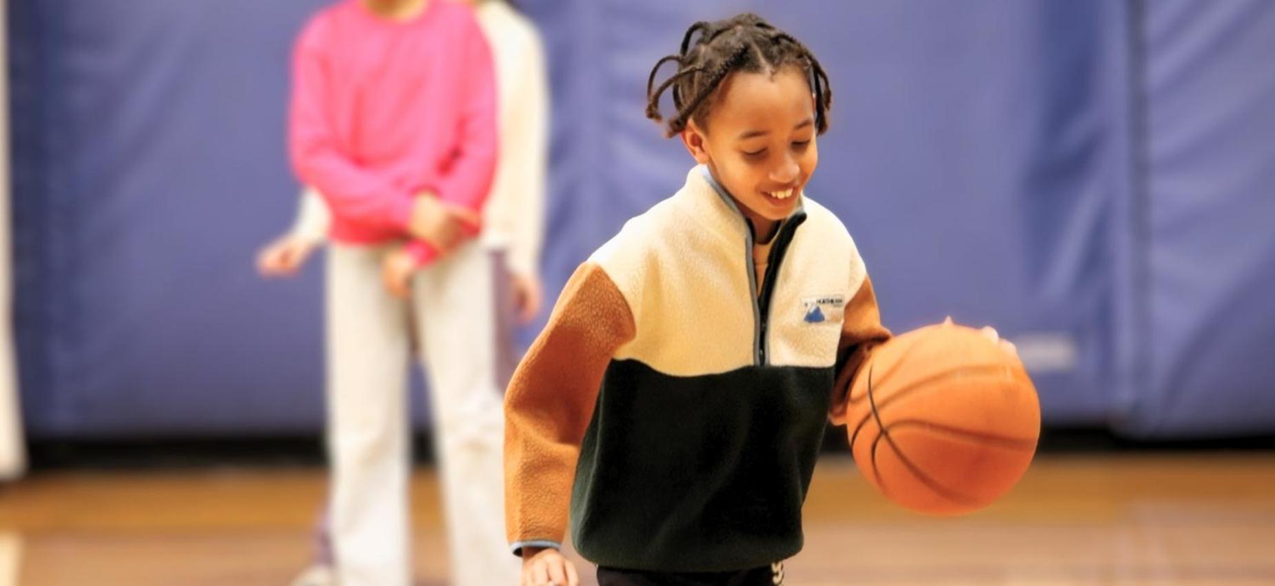 Child dribbling a basketball in a gym with other kids in the background.