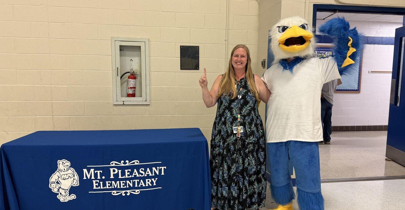 Person in a dress with a mascot in a school setting next to a table.