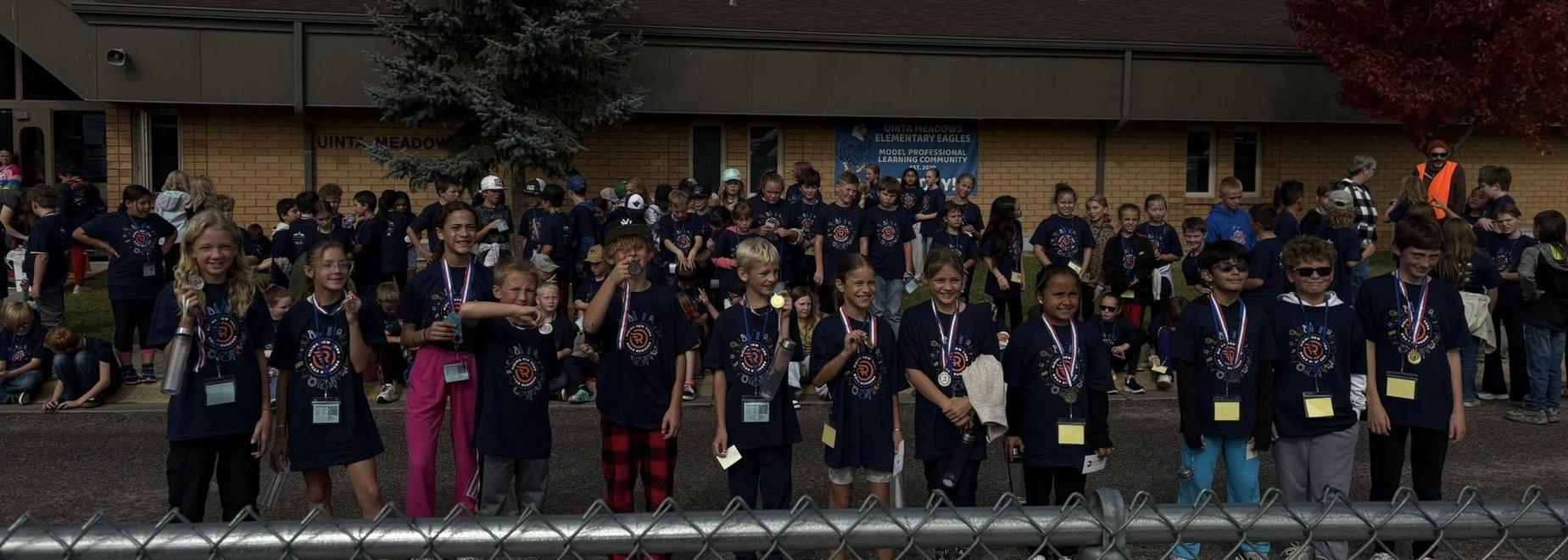 Large group of children in matching shirts at a school event with trees nearby.